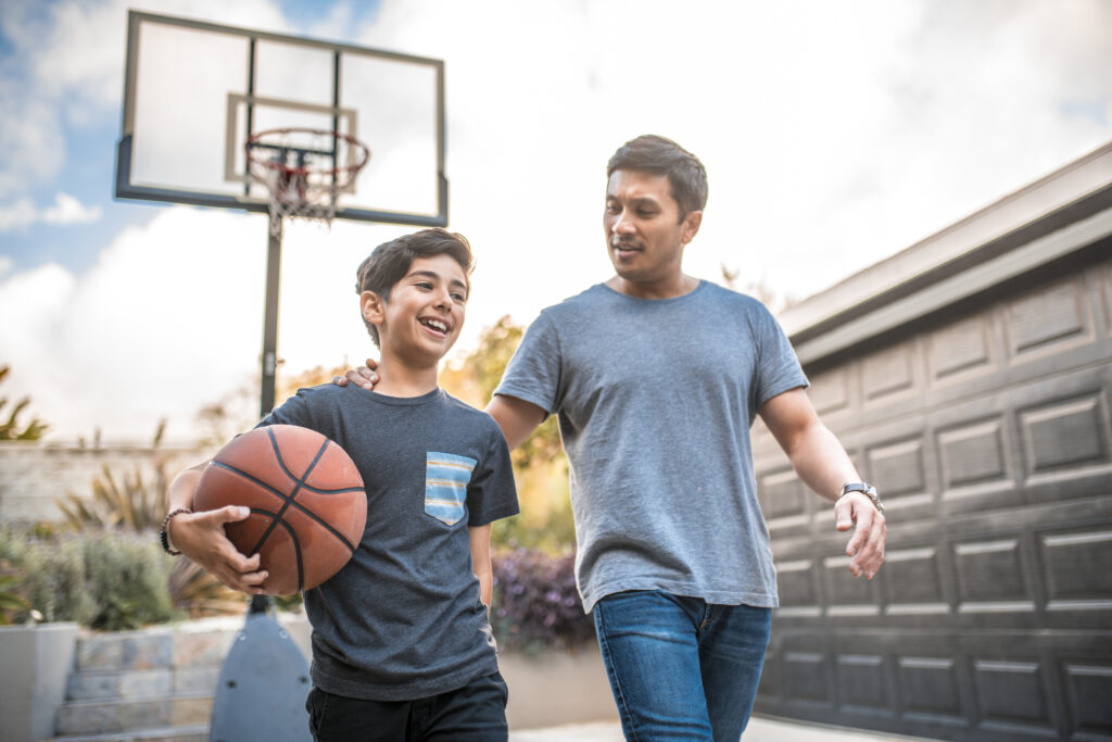 Happy boy after the basketball match with his father. Mid adult man and child are smiling in backyard. They are wearing casuals during weekend.