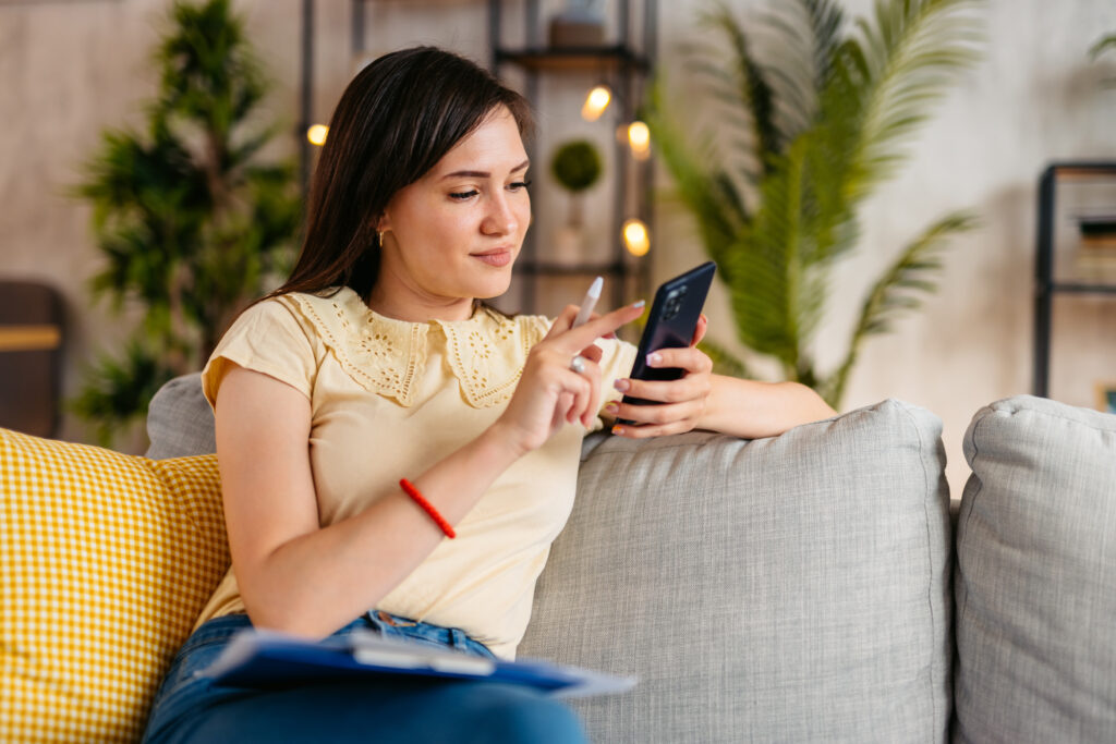 Beautiful young woman sitting on the sofa in the living room and calculating her finances on a smart phone.